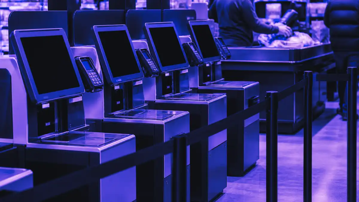 Unused self-checkout kiosks sit behind queue barriers inside a retail store while a staffed checkout lane operates in the background, showing Walmart’s shift back to cashier-led checkout.
